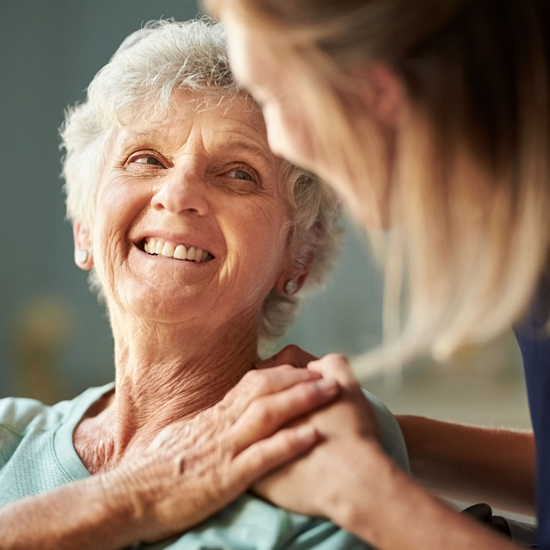 Older woman smiling as she places her hand on a younger woman's hand, which is on the older woman's shoulder.